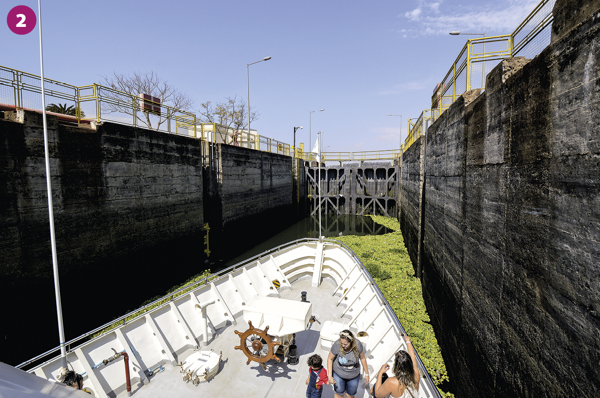Fotografia 2. Vista de embarcação pequena com algumas pessoas dentro, localizada em um canal cercado por duas estruturas verticais de concreto. Acima dessas estruturas, outras construções, feitas de ferro.