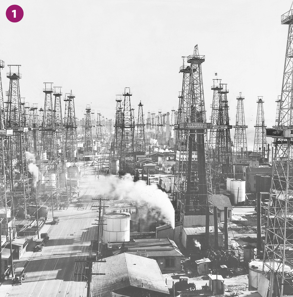 Fotografia em preto e branco 1. Vista aérea de um parque industrial, composto por diversas torres altas, tanques, chaminés soltando fumaça e vias internas de passagem de veículos.