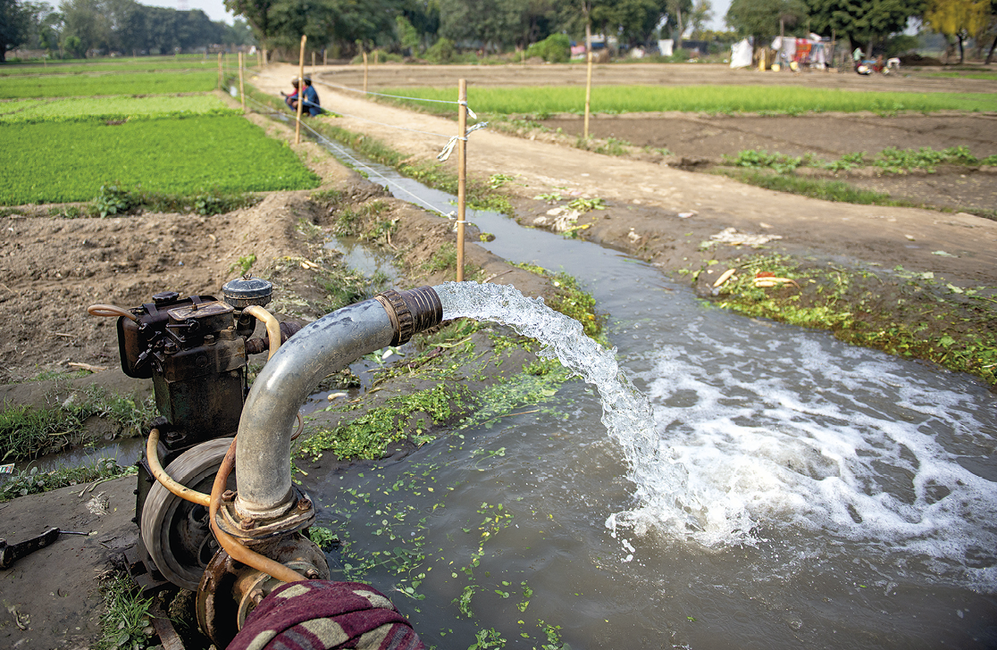 Fotografia. Uma tubulação grande despeja água em uma canaleta de irrigação. Ao redor, vegetação verde e plantações.
