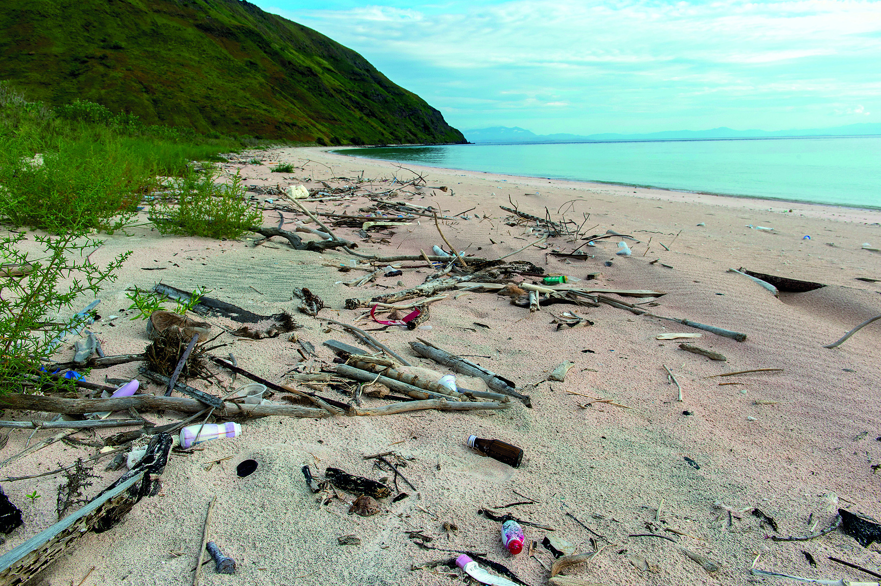 Fotografia. O ângulo da imagem evidencia em primeiro plano a faixa de areia de uma praia onde há diversos galhos de árvore quebrados e a presença de lixo e resíduos plásticos entre eles. Ao fundo, à direita, está o mar e o céu. à esquerda, vegetação arbustiva e um morro.