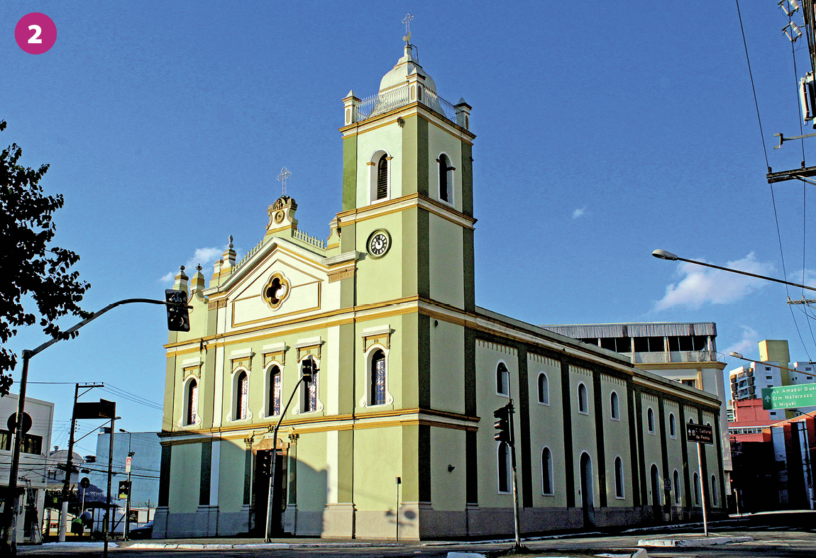 Fotografia 2. Imagem de uma igreja. As paredes são verdes com listras amarelas, e há uma estrutura retangular com uma torre e janelas com vitrais. Faróis de trânsito, vias asfaltadas, postes de luz com fiação elétrica, casas e prédios estão presentes ao redor da igreja.