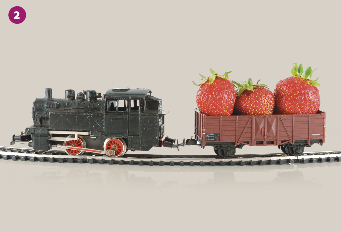 Fotografia 2. Um trem cinza em miniatura está sobre os trilhos e carrega uma carga de três morangos grandes.