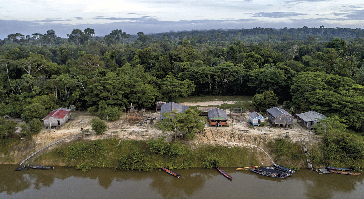 Fotografia. Vista aérea da margem de um rio. Na parte inferior está o rio de água escura e marrom, com algumas embarcações pequenas ancoradas. Logo acima, em um terreno um pouco mais elevado, estão seis construções baixas de madeira e formato retangular. Algumas delas estão erguidas do chão por meio de estacas e pilares. Ao fundo, vegetação verde e densa composta por árvores altas.