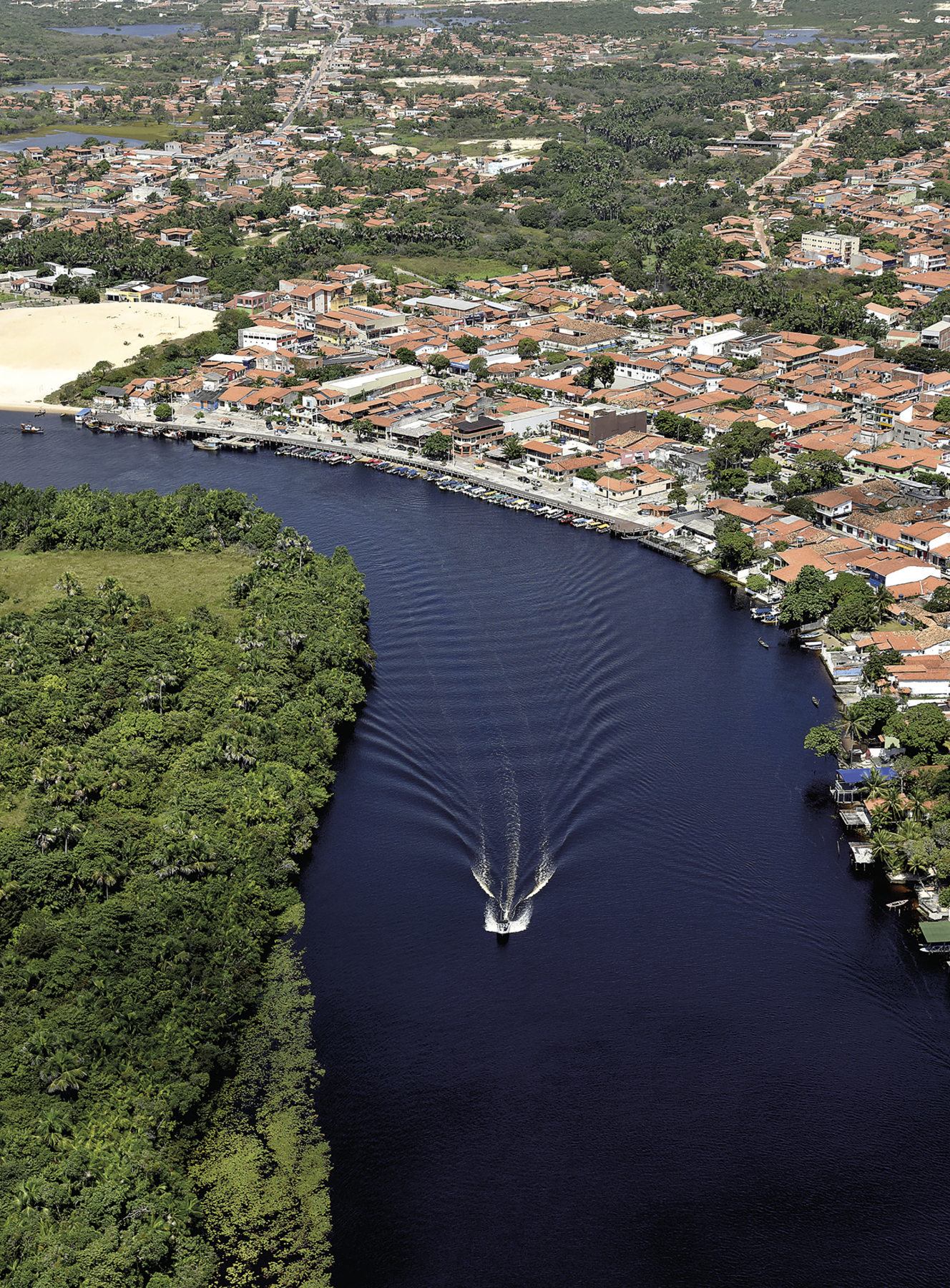 Fotografia. Vista aérea de uma cidade na beira de um rio. Ao centro está o rio de águas com tonalidade escura, ondulado por causa de uma pequena embarcação que deixou rastro ao se movimentar. Ao lado esquerdo do rio, há a presença de densa vegetação verde. Ao lado direito, grande concentração de construções baixas, sobretudo casas, além de área asfaltada e barcos ancorados na margem do rio.