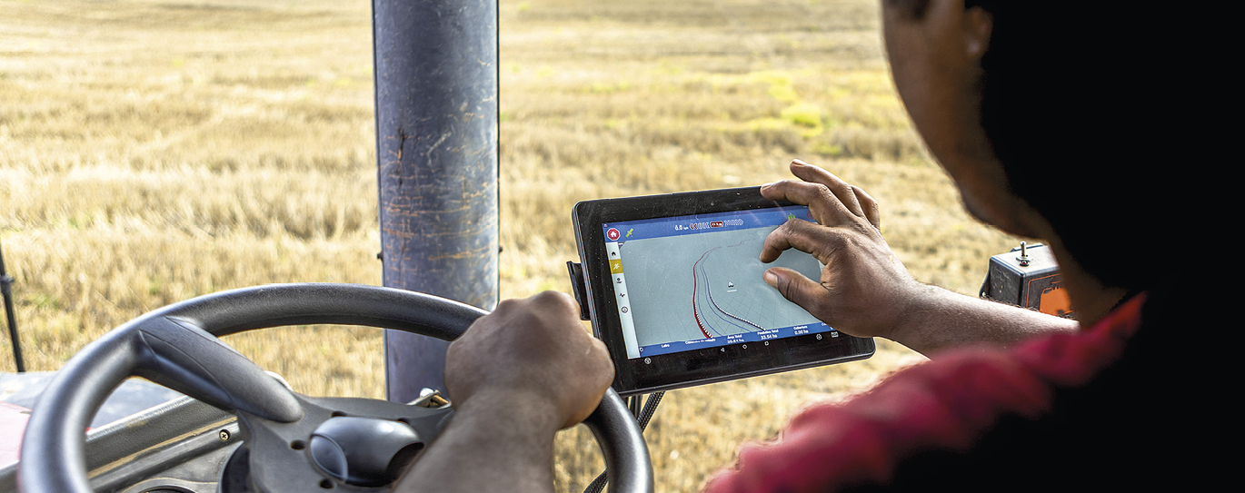 Fotografia. Uma pessoa dirige um maquinário agrícola. Para se orientar, utiliza um aparelho GPS. Uma de duas mãos está sobre o volante, e a outra sobre a tela do aparelho.
