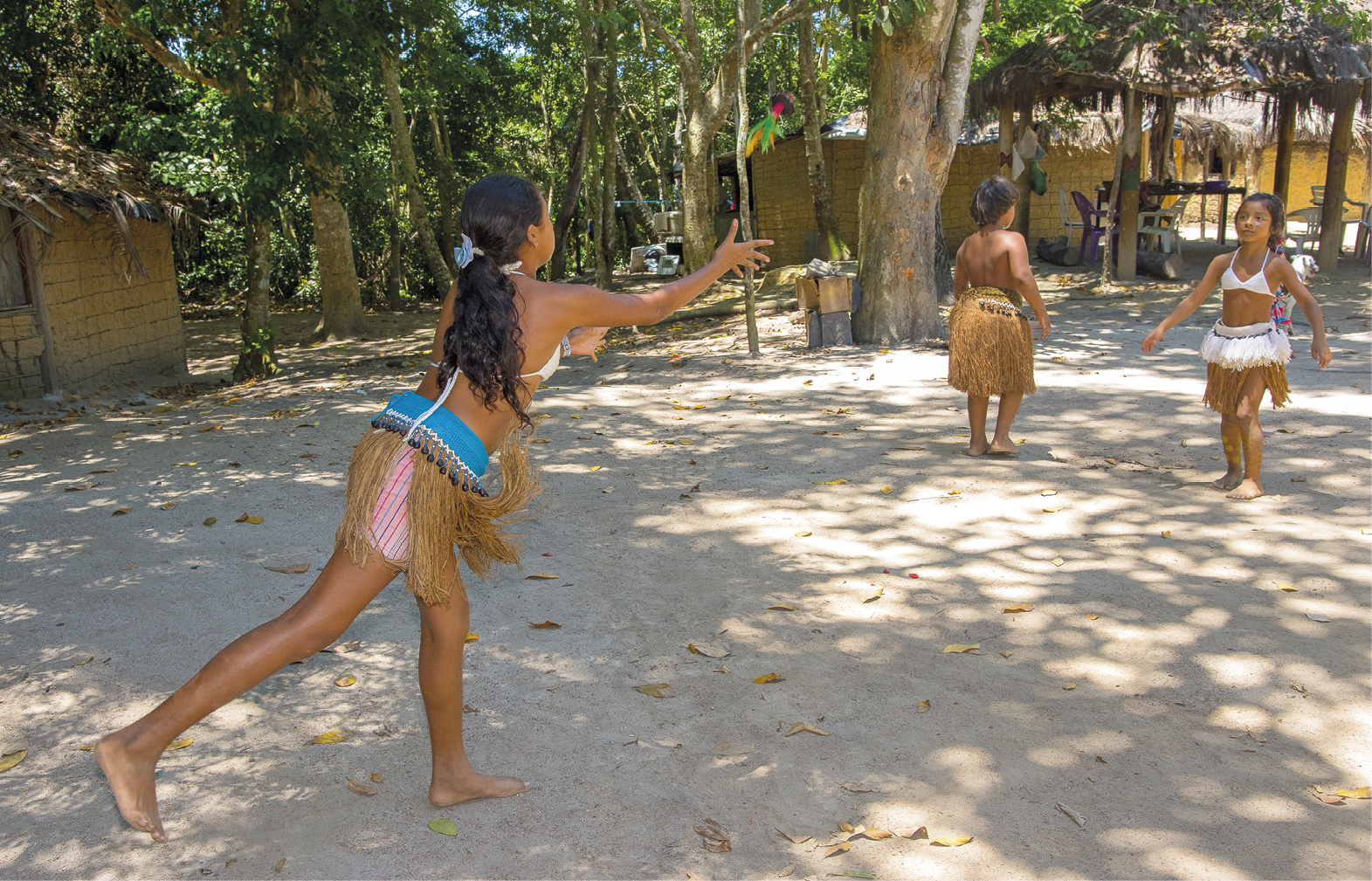 Fotografia. Duas crianças indígenas brincam de peteca em um pátio, enquanto outra aparece mais ao fundo. O chão é de terra batida. Em segundo plano, há construções entre árvores com paredes de pau a pique.