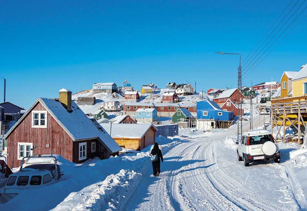 Fotografia. Vista de um caminho largo e coberto por neve ao centro, onde uma pessoa agasalhada caminha. As casas à beira desse caminho são coloridas, revestidas de madeira, e possuem o telhado íngreme. Acima, o céu está azul e sem nuvens.