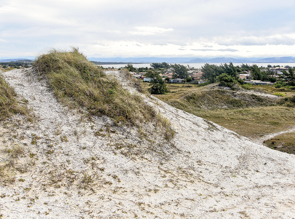 Fotografia. Em primeiro plano está um pequeno morro de coloração esbranquiçada, com vegetação rala e rasteira em seu topo. Ao fundo, vegetação rasteira e algumas árvores e construções.