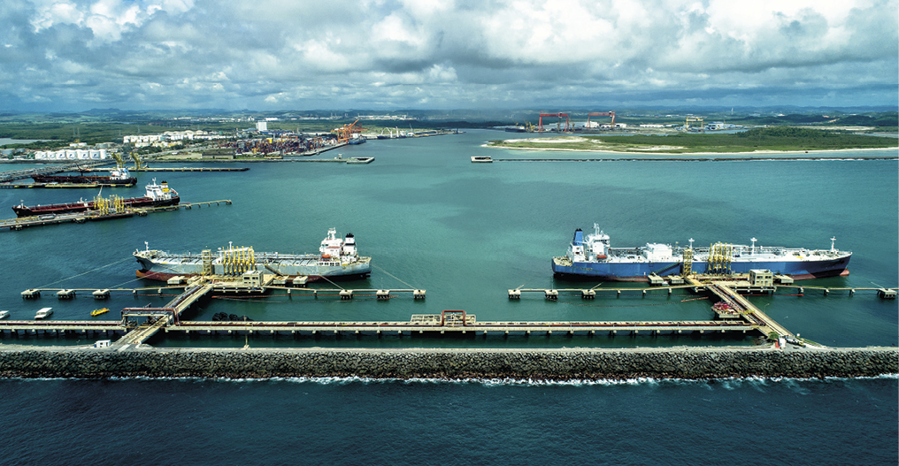 Fotografia. Vista aérea de uma zona portuária onde, em primeiro plano, dois grandes navios estão ancorados próximos à construção em forma de barreira, chamada de quebra-mar. O quebra-mar é composto por paredes de rochas que impedem que a área do porto seja afetada por ondas. As águas do mar são verdes e transparentes. Ao fundo, no canto direito, há uma pequena mancha verde indicando a vegetação do local.