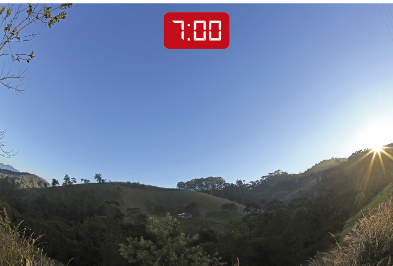 Fotografia. Vista de uma uma paisagem com declives, gramado verde e algumas árvores na parte inferior. Na parte superior, o céu está azul e o Sol desponta por trás de um morro ao lado direito. A paisagem apresenta apenas alguns pontos iluminados pela luz solar, permanecendo a maioria com sombras. No topo da imagem, um visor de relógio digital indica o horário: 7 horas.