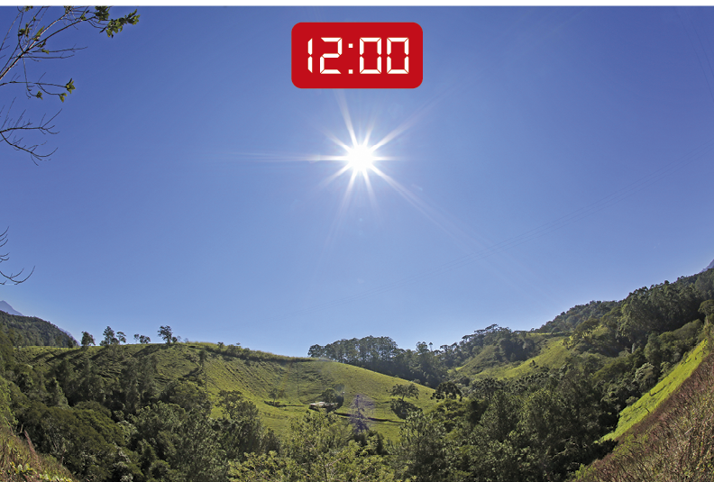 Fotografia. Vista de uma uma paisagem com declives, gramado verde e algumas árvores na parte inferior. Na parte superior, o sol está posicionado no centro do céu azul. A vegetação e o gramado estão completamente iluminados pela luz solar e não há áreas de sombras. No topo da imagem, um visor de relógio digital indica o horário: 12 horas.