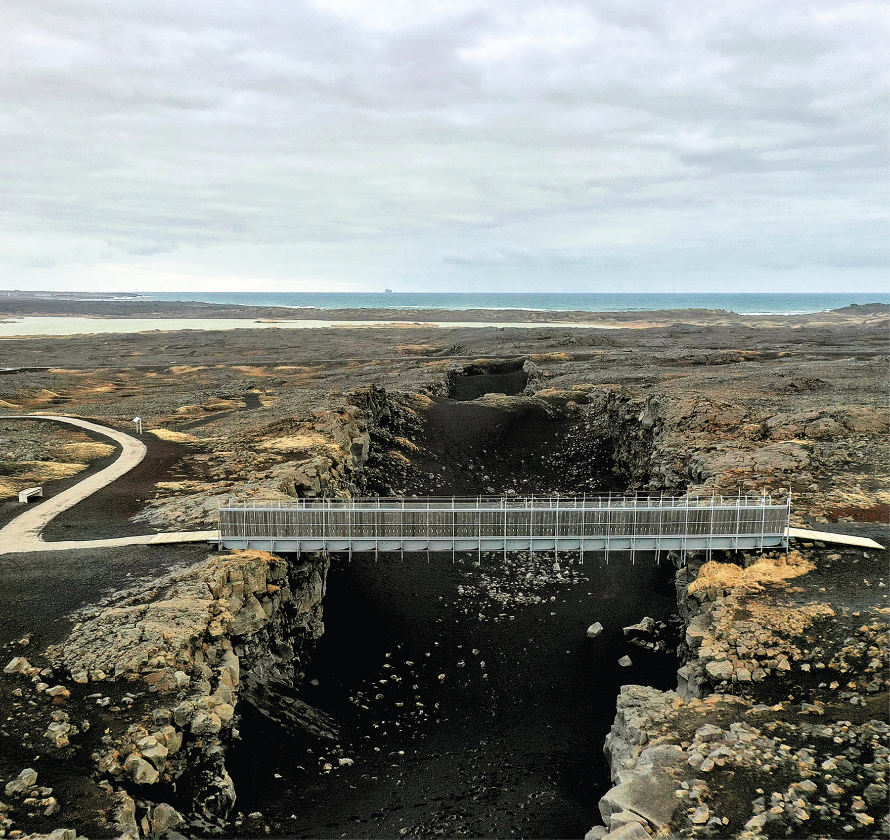 Fotografia. Vista de um vale descampado e rochoso, onde ao centro há uma fenda escura. Uma ponte cinza liga as extremidades dessa fenda e se conecta a uma estrada localizada na parte esquerda da imagem. Ao fundo está o mar e o céu acima está cinza e nublado.