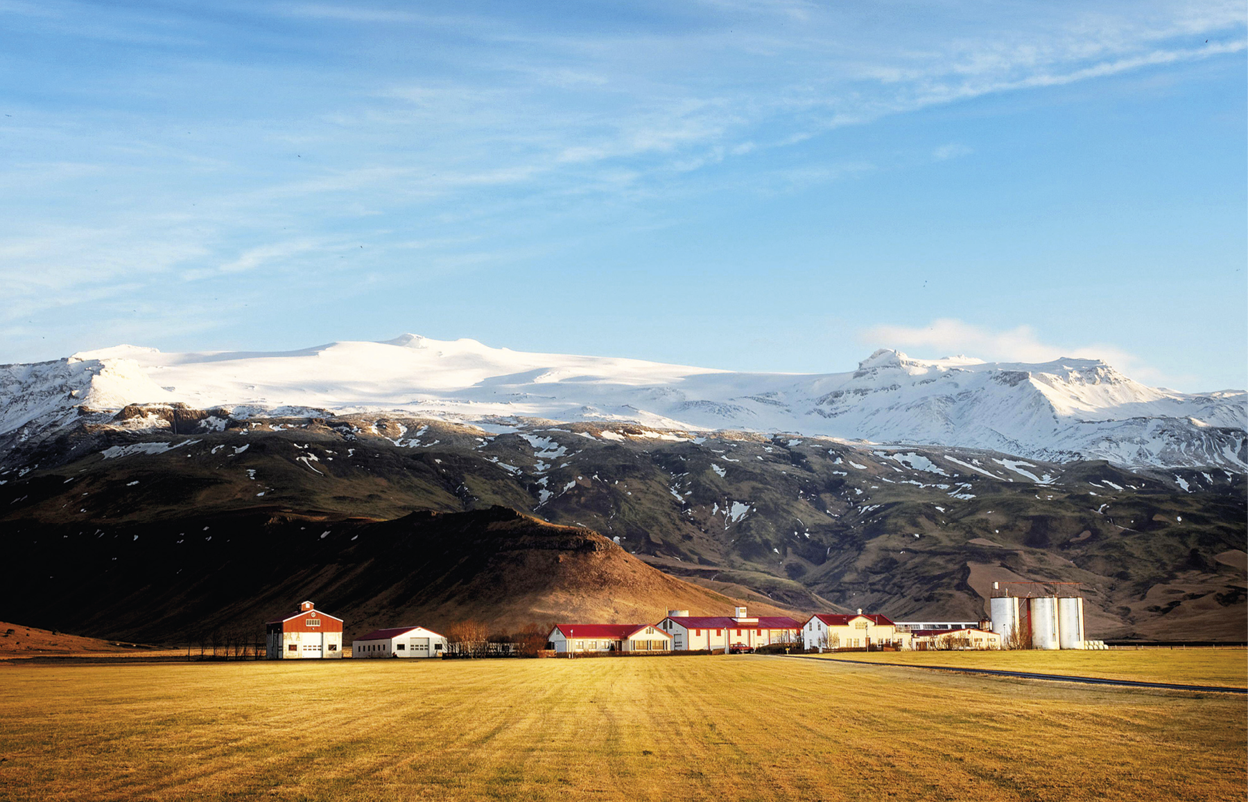 Fotografia. Em primeiro plano extenso campo de coloração amarelada. Em segundo plano, algumas construções com o telhado vermelho, além de torres arredondados à direita. Ao fundo, cadeia de montanhas em tons amarronzados e cume com neve. Acima, o céu está azul e a paisagem é iluminada pela luz do sol.