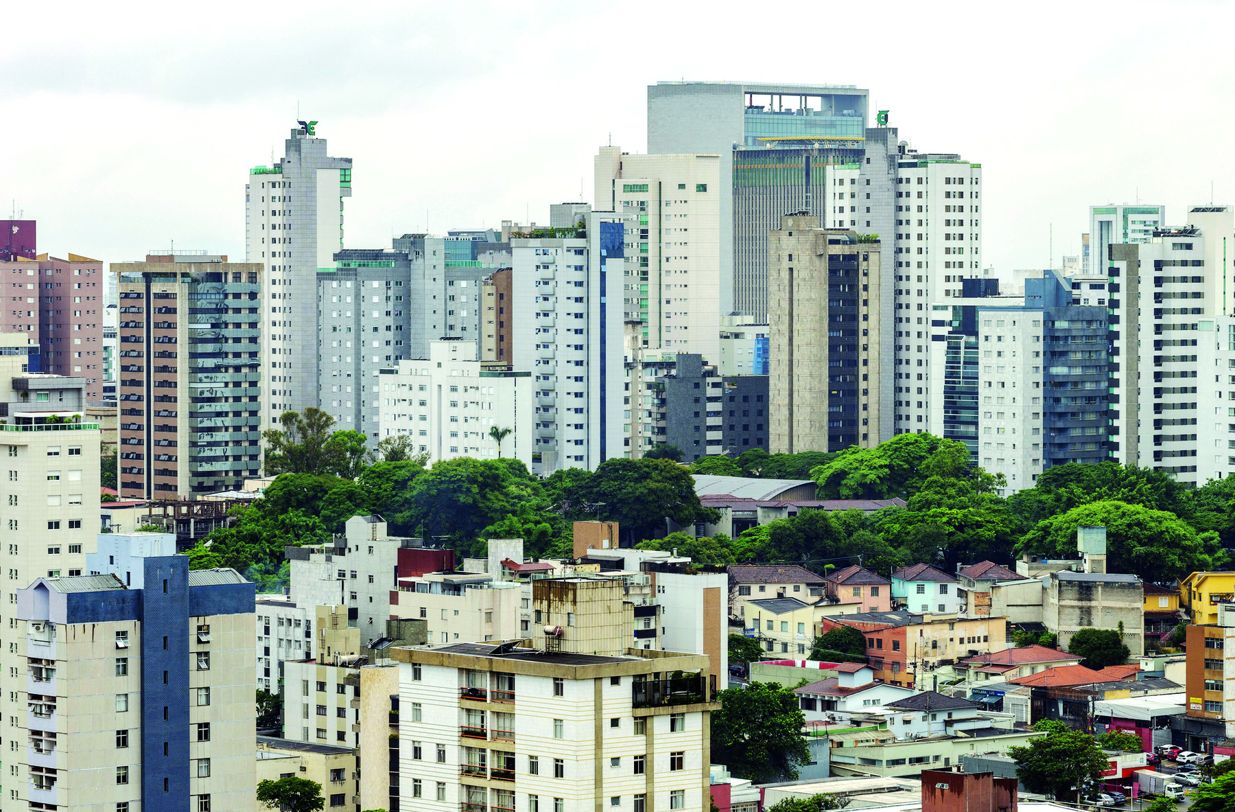 Fotografia. Vista de uma concentração de prédios e casas. Na parte superior o céu está nublado. Ao centro há a presença de uma área arborizada.