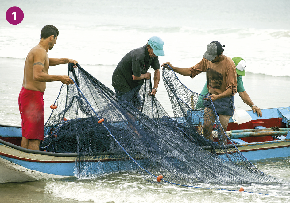 Fotografia 1. Quatro homens em pé sobre uma canoa de cor azul. Três deles seguram uma rede de pesca com boias laranjas em suas extremidades. O quarto homem está atrás dos demais e se apoia em uma das beiradas do barco. Ao fundo está o mar.
