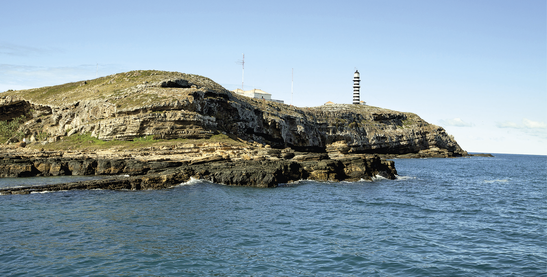 Fotografia. No primeiro plano, o mar com águas em tons de azul. No segundo plano, é possível avistar um rochedo em tons de cinza e verde, devido à cobertura vegetal rasteira em alguns trechos. Ao fundo, na parte direita do rochedo, há um farol e poucas construções. A paisagem foi fotografada durante o dia e o céu está claro.