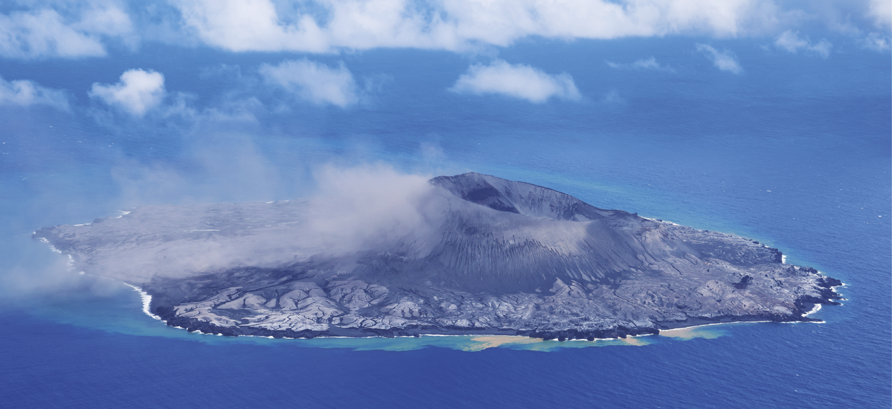 Fotografia. Vista aérea de uma ilha cujo centro é ocupado por um vulcão com a superfície rochosa em tons de cinza escuro. O centro do vulcão possui uma grande cavidade arredondada. Ao redor do vulcão, o terreno apresenta um tom de cinza mais claro. Ao redor da ilha há água em tons de azul. O céu, igualmente azul, apresenta algumas nuvens.