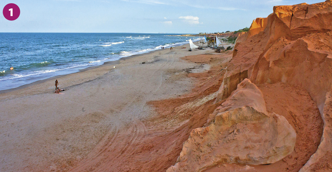 Fotografia 1. Vista do mar e de uma praia, à esquerda, e de um terreno elevado, irregular e em tons de marrom, à direita.