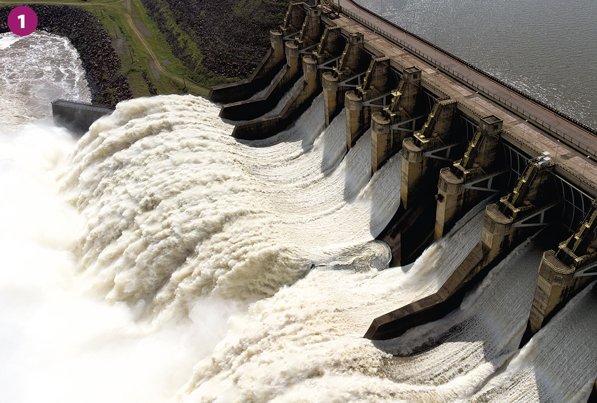 Fotografia 1. Vista de uma barragem. Grandes quantidades de água atravessam estruturas de concreto e metal, em um movimento de queda que faz com que a água tenha aparência branca.