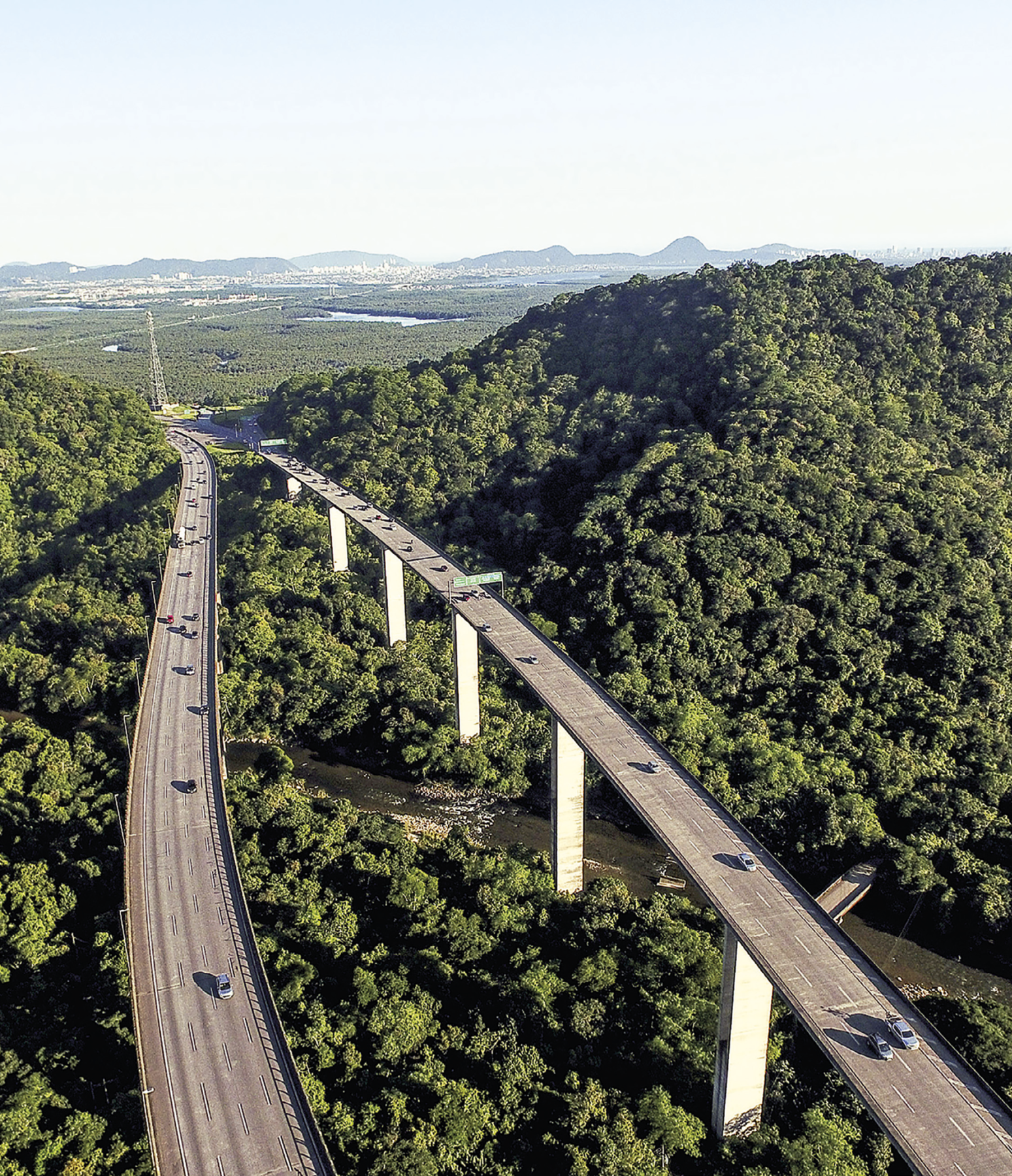 Fotografia. Vista aérea de uma rodovia, cujo primeiro plano apresenta duas estradas erguidas com pilares de concreto. Nela há diversos carros que seguem o mesmo sentido, do canto superior da imagem para o canto inferior. A rodovia ergue-se sobre uma densa vegetação com predominância de árvores e um rio, elementos que se encontram no segundo plano da imagem. No terceiro plano, há um terreno ocupado principalmente por árvores, rios e a continuidade da estrada. No quarto plano, está a paisagem de uma cidade e diversos morros.