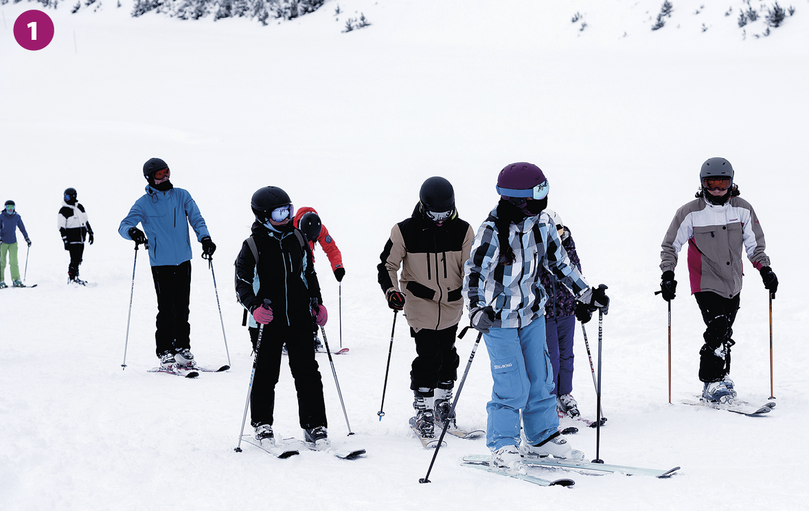Fotografia. Diversas pessoas vestindo calça, casaco, gorro e máscara estão em pé sobre equipamento de esqui, segurando bastões, em área coberta de neve.