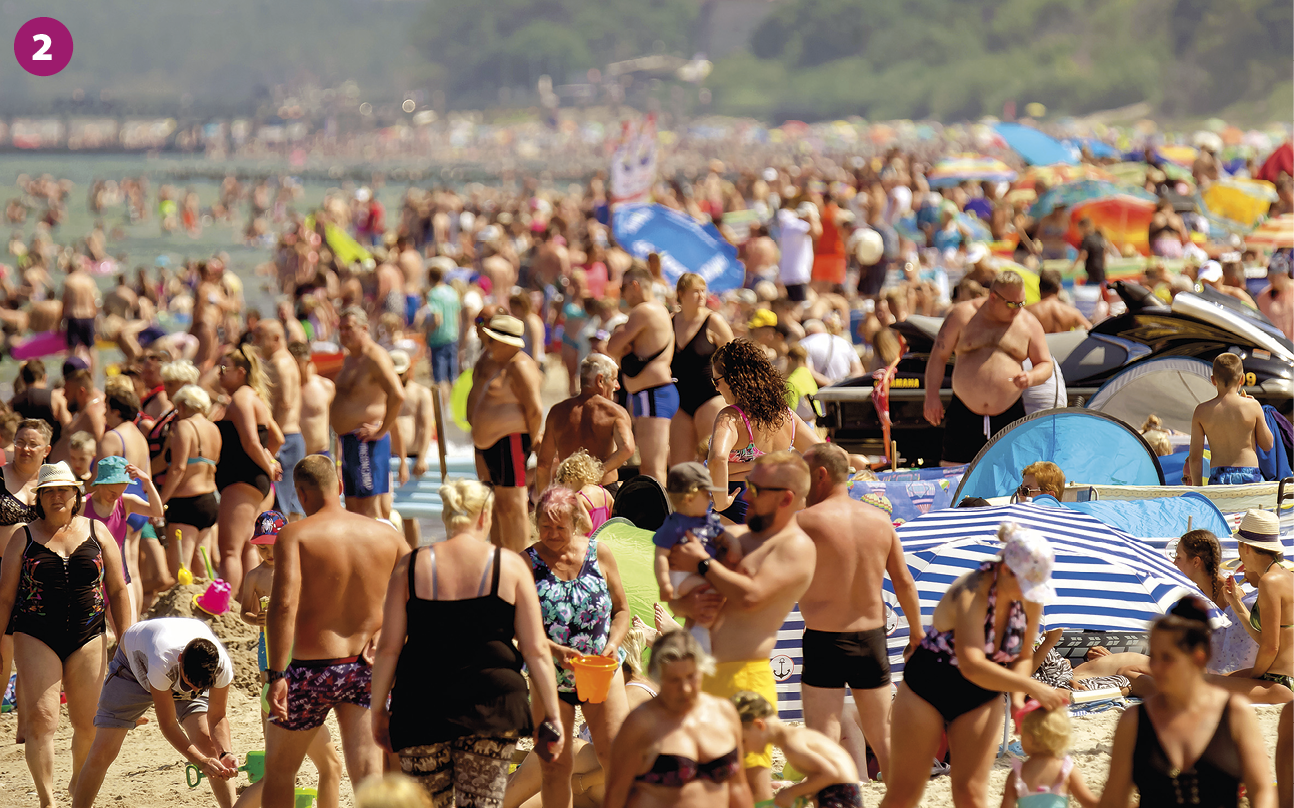 Fotografia. Diversas pessoas vestindo roupas de banho, como sungas, maiôs, biquínis e chapéus, estão em pé na areia da praia. Entre as pessoas, há toalhas, barracas e guarda-sóis. Ao fundo, presença de pessoas no mar.