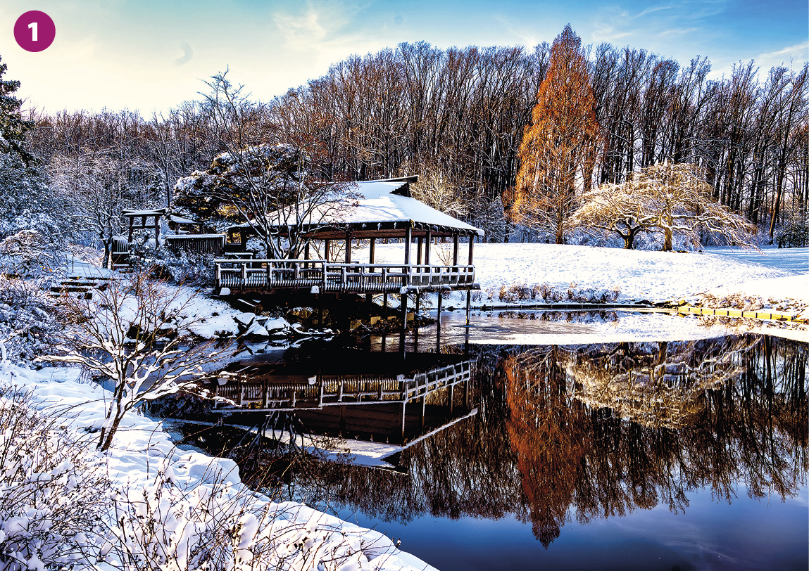 Fotografia 1. No primeiro plano, um corpo d'água cristalino com margens cobertas por neve e vegetação rasteira seca. No segundo plano, uma casa de madeira na beira do corpo d'água e refletida na água.  No terceiro plano, floresta densa com vegetação arbórea de grande porte, folhas secas, amareladas e refletidas na água. Acima, o céu azul.