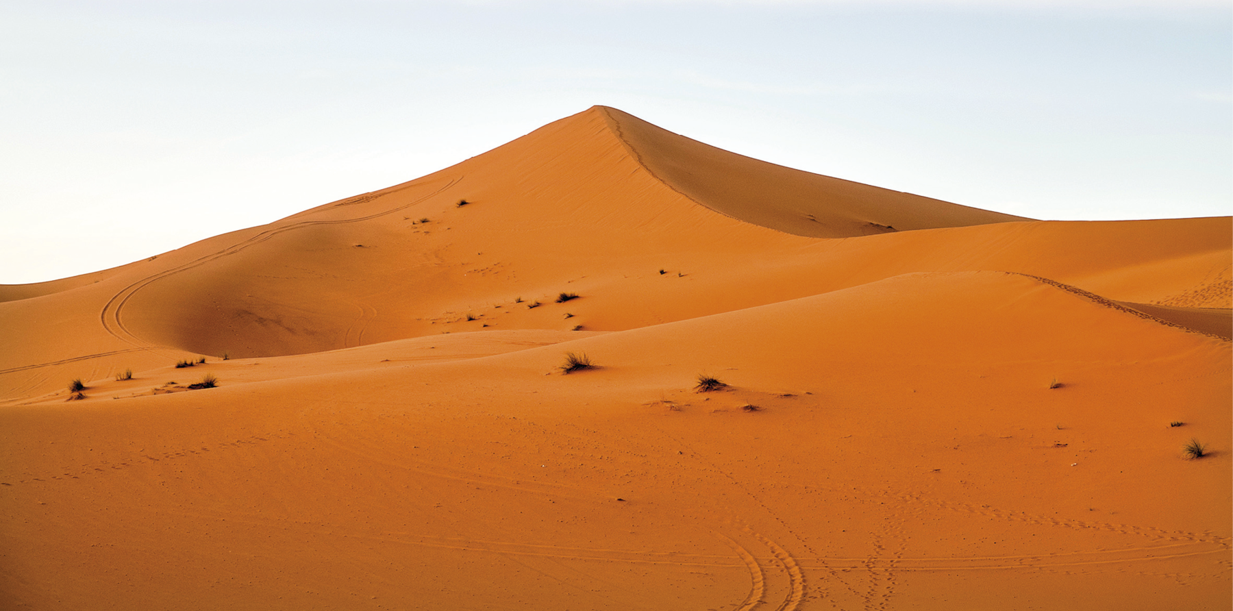 Fotografia. Vista panorâmica de uma duna alaranjada no deserto. Acima, o céu em tons de azul.