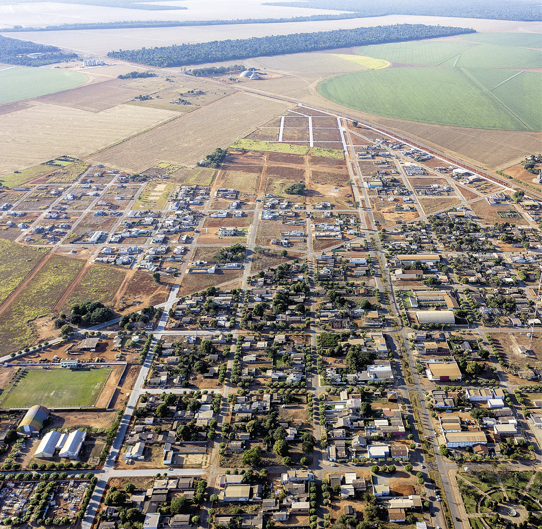 Fotografia. Vista aérea de uma cidade pouco adensada, composta por um traçado de ruas paralelas e perpendiculares e quarteirões com casas e árvores. Entre os quarteirões, ruas asfaltadas com alguns veículos. À esquerda, há galpões brancos e um campo de futebol. Ao fundo, fragmento florestal denso e propriedades agrícolas de diversos tamanhos e estágios de cultivos distintos.