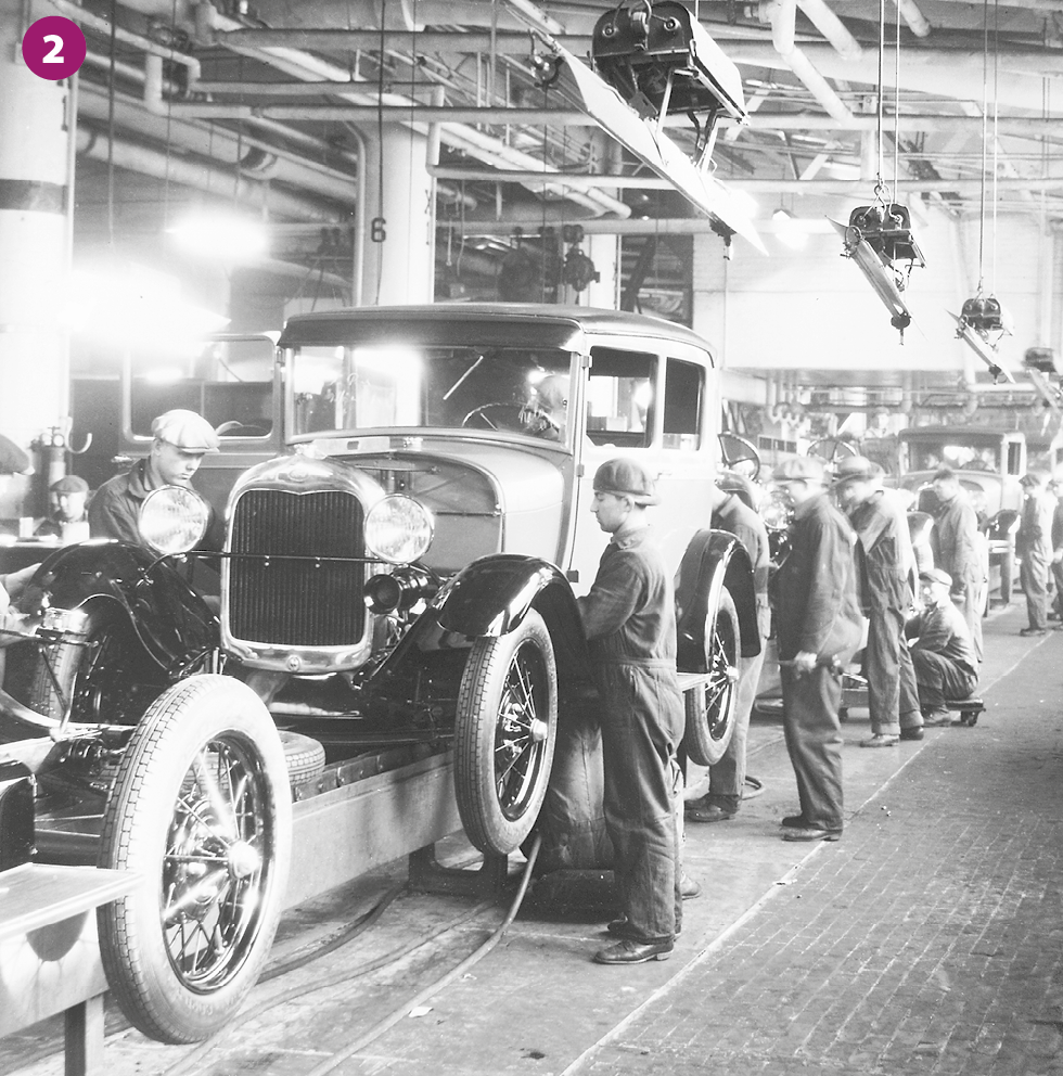 Fotografia em preto e branco 2. Vista para uma linha de produção de automóveis, com diversos veículos antigos enfileirados e em cima de um trilho. Nas laterais, homens manuseiam peças de montagem junto aos automóveis. Acima, o teto com o encanamento aparente, lustres e lâmpadas.