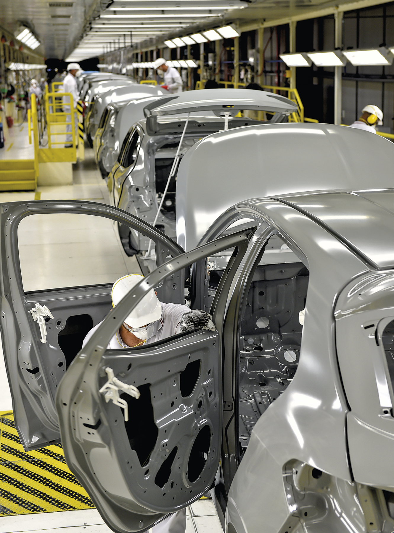 Fotografia. Vista para uma linha de produção industrial com automóveis enfileirados. No primeiro plano, um veículo prateado sendo montado, com as portas abertas e sem as peças internas. Ao lado da porta esquerda dianteira, um homem usando capacete, máscara e uniforme branco está agachado. No segundo plano, outros veículos enfileirados. À esquerda, escada com corrimão amarelo.