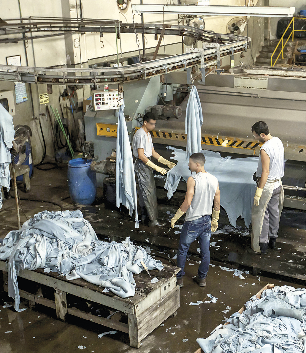 Fotografia. Vista para um ambiente fechado com diversos equipamentos e máquinas industriais. Entre as máquinas, três homens em pé, usando camiseta e calça, estão ao redor de uma bancada uma peça em tom azul claro. À frente, duas bancadas com uma pilha de peças do mesmo tom. Outras peças estão penduradas em um suporte instalado na parte superior do local.