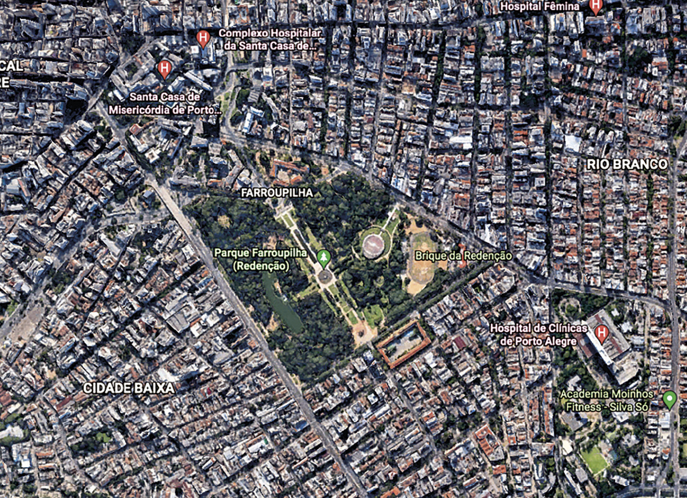 Imagem de satélite. Vista vertical de uma cidade, com uma grande área verde na porção central, rodeada de quarteirões densamente ocupados por edificações, ruas e avenidas. Na porção direita inferior, presença de grandes galpões rodeados de áreas verdes.