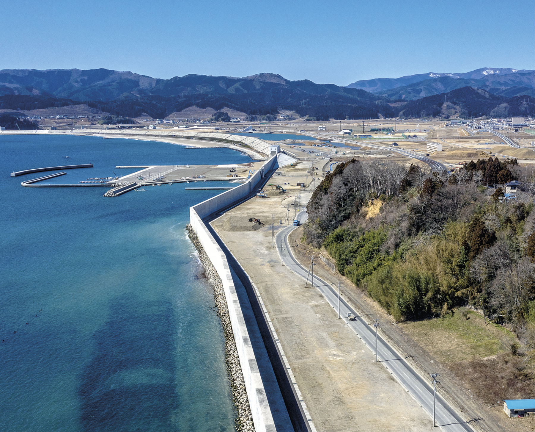 Fotografia. Vista de um muro branco e extenso ao centro. À esquerda está o mar com águas verdes e claras e, à direita, uma estrada se estende desde o primeiro plano até o fundo. Há uma porção de vegetação verde concentrada do lado direito da fotografia. O segundo plano é cortado por algumas estradas e outras construções humanas. Ao fundo, uma cadeia de morros cobertos por vegetação verde.