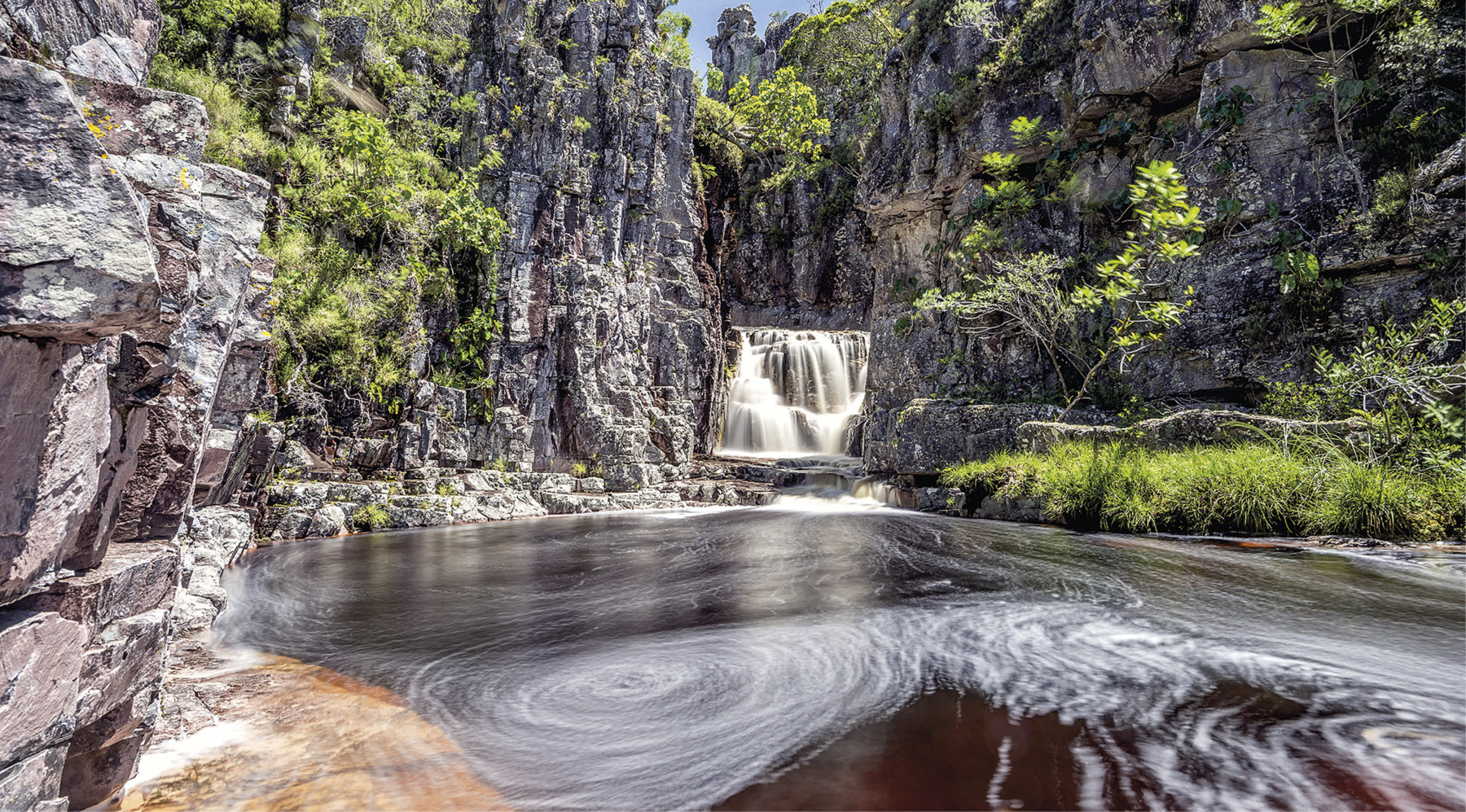 Fotografia. Em destaque, uma cachoeira cercada por paredões rochosos e vegetação verde. No primeiro plano, há uma grande quantidade de água que deriva da cachoeira.