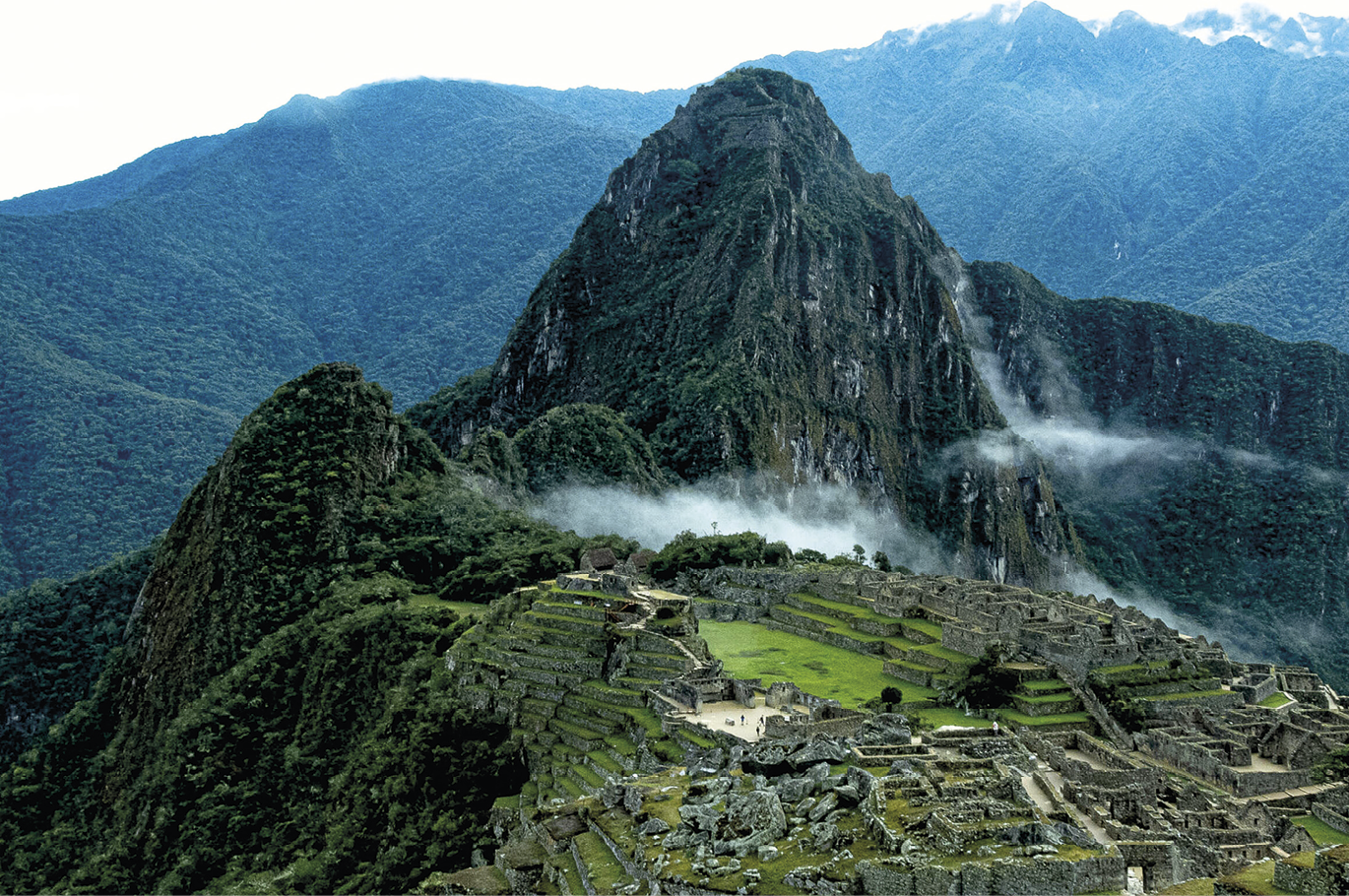 Fotografia. Vista aérea das ruínas de uma cidade, onde predominam construções feitas de pedras em tons acinzentados, muitas delas cobertas por vegetação, como grama e musgo. Esses elementos estão no primeiro plano da fotografia. No segundo plano, há uma área montanhosa, na qual se destaca uma formação rochosa ao centro da imagem, mais alta que as demais e com formato pontiagudo. Há ainda a presença de neblina que se aproxima da elevação montanhosa. No terceiro plano, é possível observar mais montanhas.