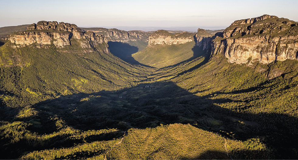 Fotografia. Em primeiro plano, há um terreno coberto por vegetação densa e verde. Essa vegetação possui declives de ambos os lados, que levam às formações rochosas de planaltos que se localizam no segundo plano. Os planaltos estão tanto do lado direito como do esquerdo da fotografia e se estendem até o fundo da imagem. Os planaltos apresentam cobertura vegetal em suas superfícies e, por isso, possuem coloração verde nesta parte, enquanto as suas escarpas têm tons terrosos.