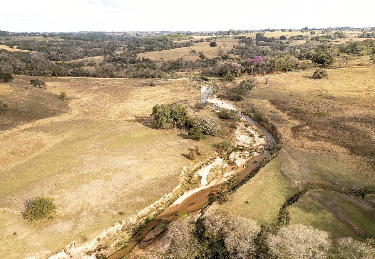 Fotografia. Vista de uma terra seca em tons de marrom claro e verde. No centro há a passagem de um rio com pouco volume de água e uma material semelhante à areia em uma das margens. Ao fundo, algumas árvores e vegetação baixa. Acima, o céu com nuvens.