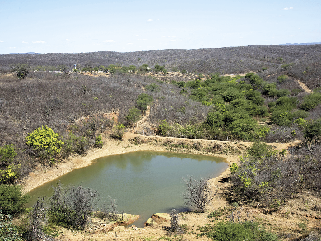 Fotografia. Vista de um rio cercado por uma pequena faixa de terras secas. Ao redor há vegetação arbustiva e seca, com poucas árvores. Acima, o céu em tons de azul.