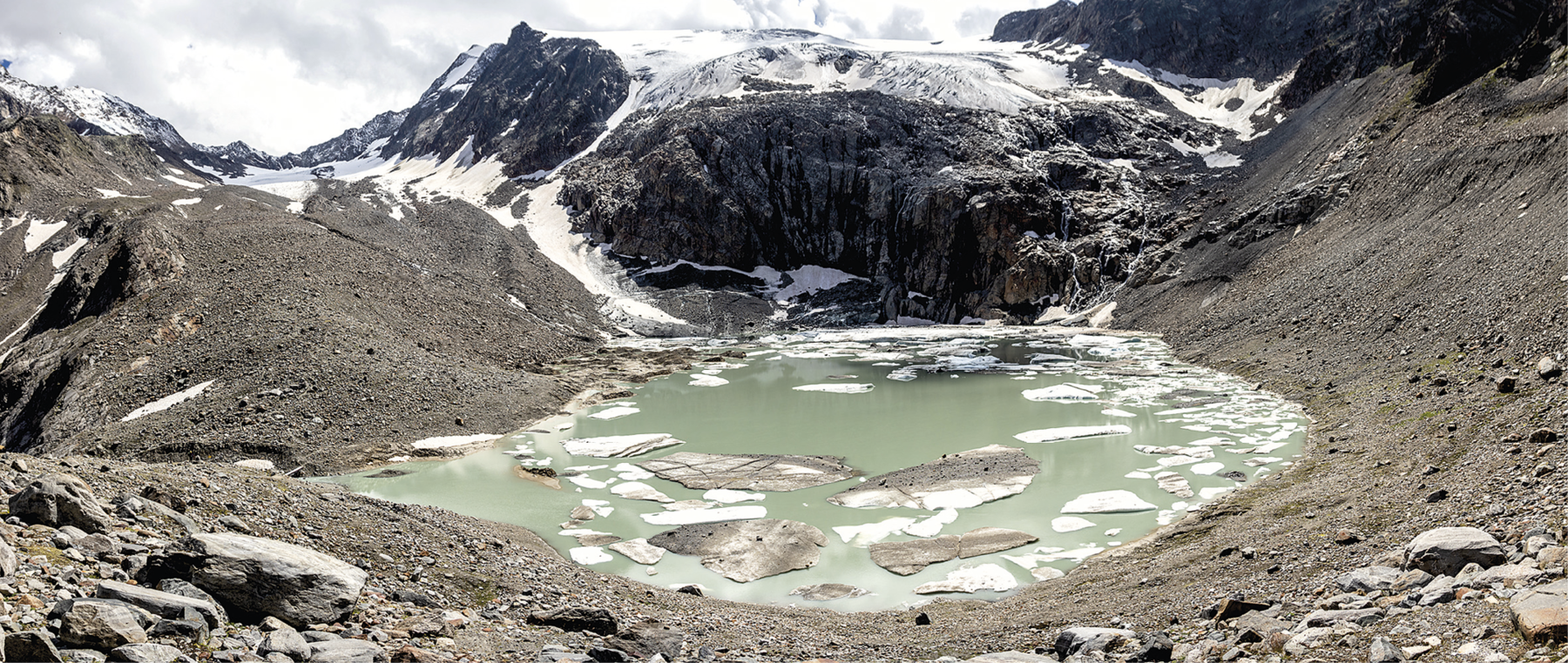 Fotografia. Vista de um lago congelado com blocos de gelo na superfície da água. Ao redor há uma rocha e terras em tons escuros. Ao fundo, um terrenos bastante elevado com neve no topo.