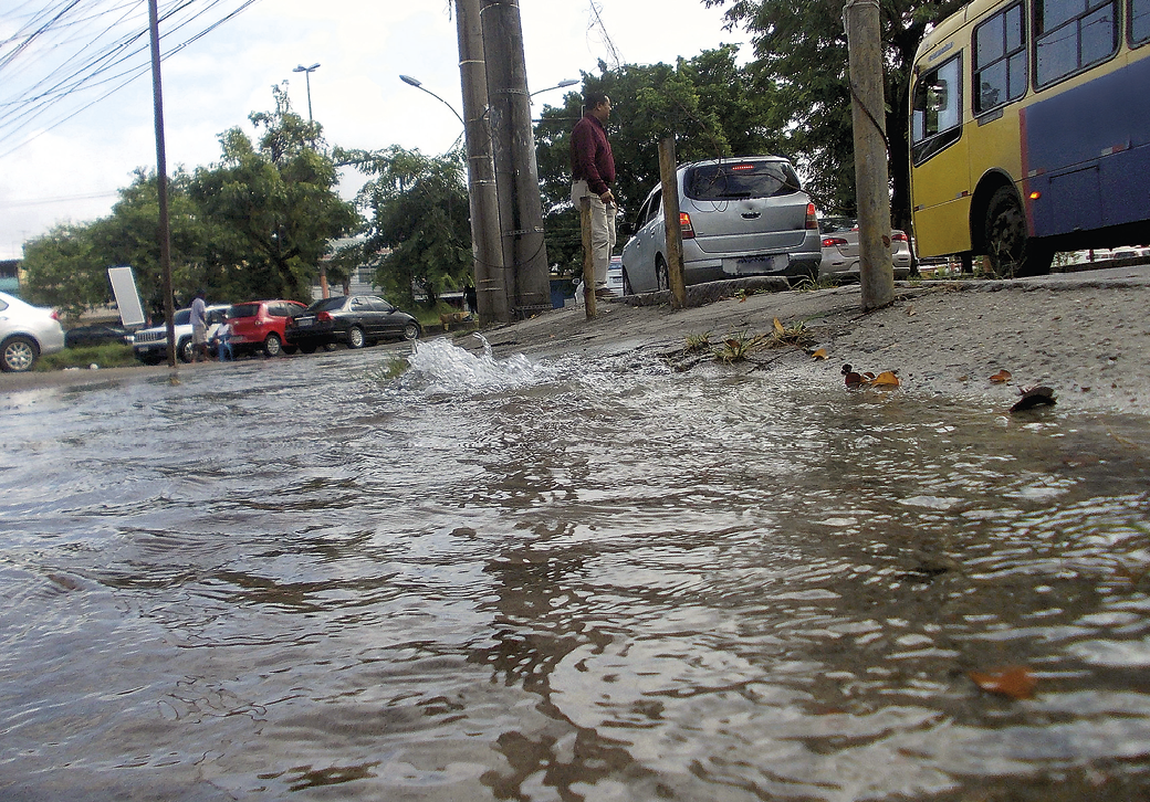 Fotografia. Rua vista por um ângulo perto do chão. O lugar está inundado. Ao lado há terra, postes e carros na rua. Ao fundo, árvores.