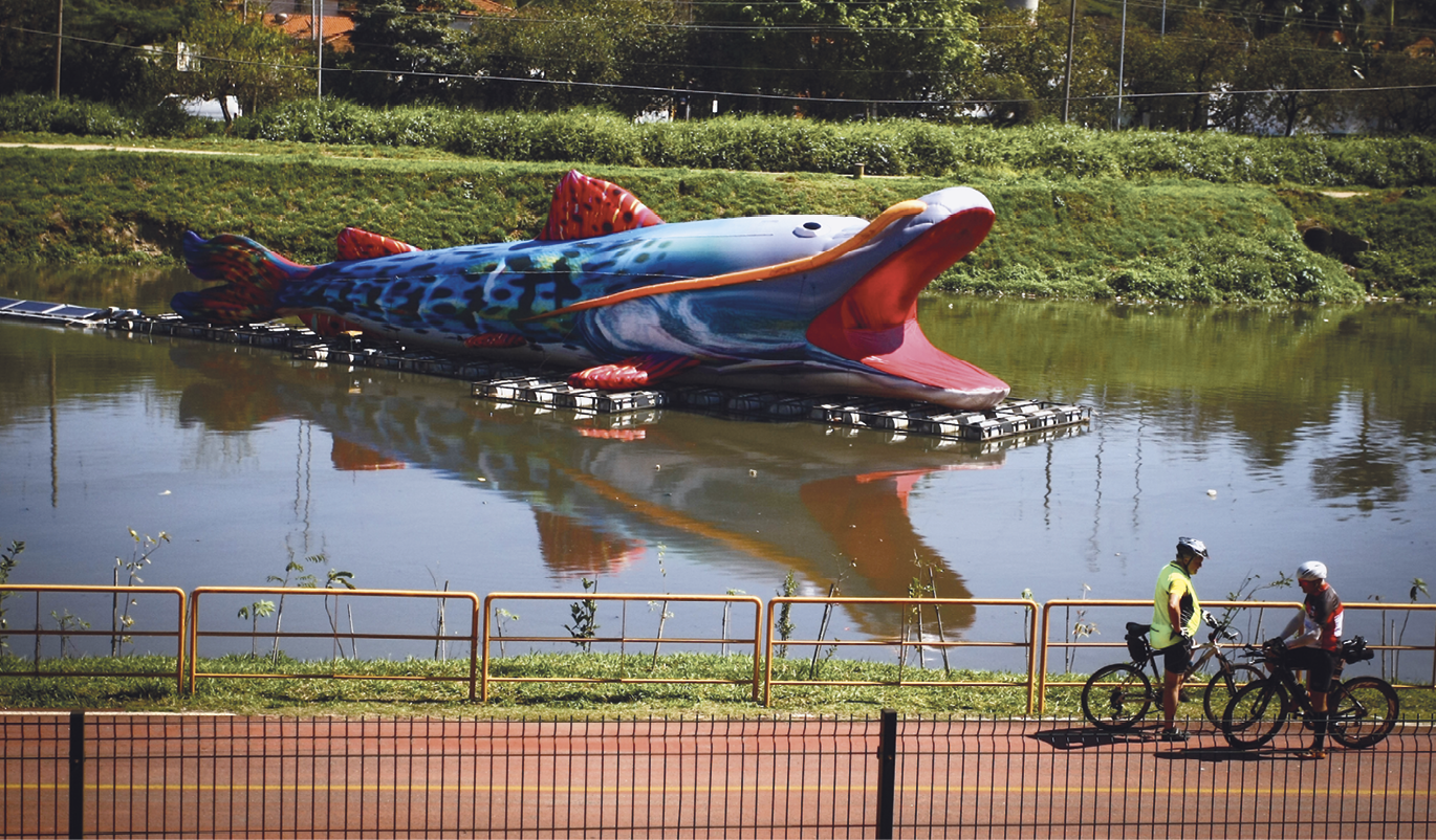 Fotografia. Ao centro, um peixe grande e colorido de borracha pintado de azul e vermelho, com a boca aberta, está sobre as águas de um rio. Na parte de baixo da imagem, em uma ciclovia, dois ciclistas. Ao fundo, área verde com a vegetação baixa.