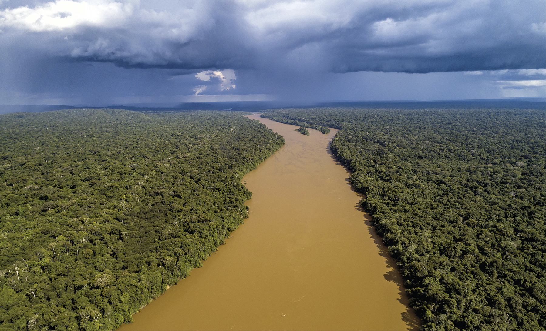 Fotografia. Vista aérea de uma vasta área plana. No centro, um rio extenso e de água marrom. Nas laterais, uma grande floresta densa e verde nas margens do rio. Acima, o céu com nuvens cinzas e carregadas.