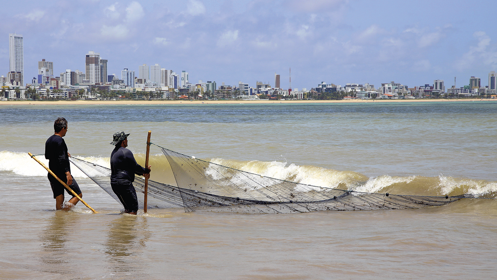 Fotografia. No primeiro plano, dois homens seguram uma rede extensa de pesca dentro do mar, com a água na altura do joelho. No segundo plano, faixa de areia e edifícios e casas na orla da praia.