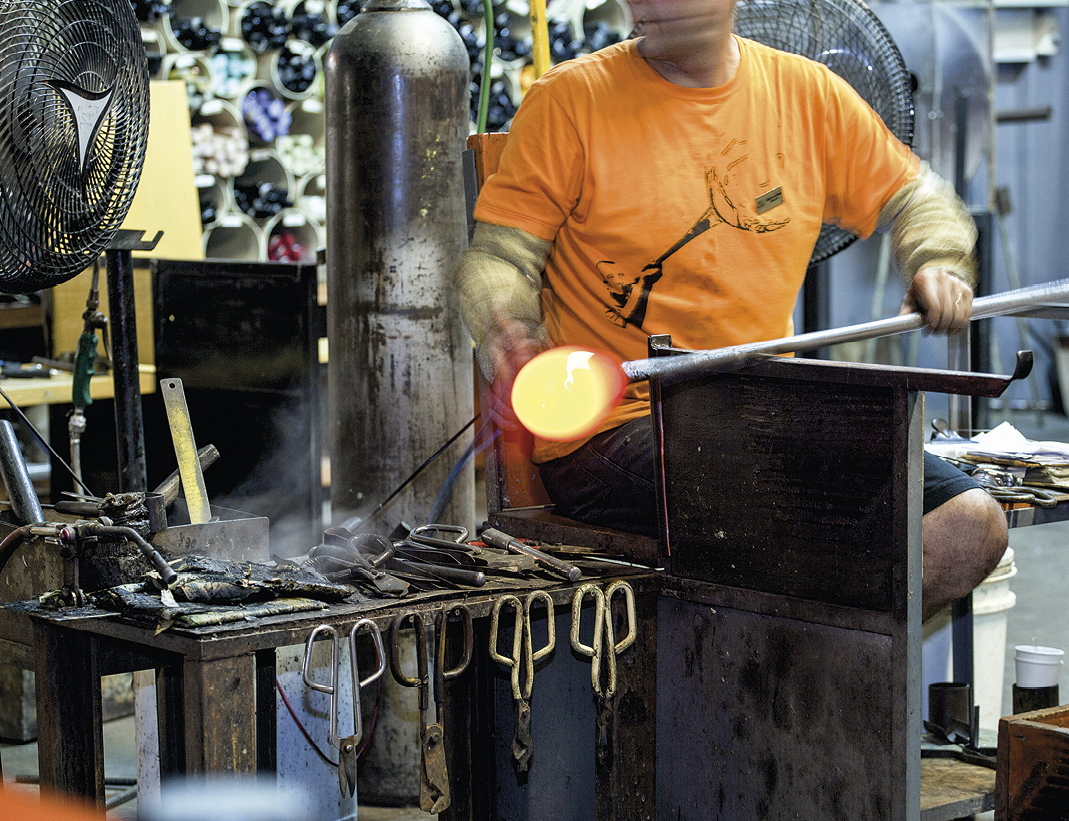 Fotografia. Destaque para um homem de camiseta laranja, sentado e manuseando um bastão fino com uma ponta de metal fundido arredondado e de tons de amarelo e vermelho. Abaixo do bastão, há um suporte que mantem o objeto na horizontal. Ao lado do homem, há uma mesa com diversas peça, como tesoura e um grande cilindro de metal. Ao fundo, diversos equipamentos industriais e suportes de armazenamento.