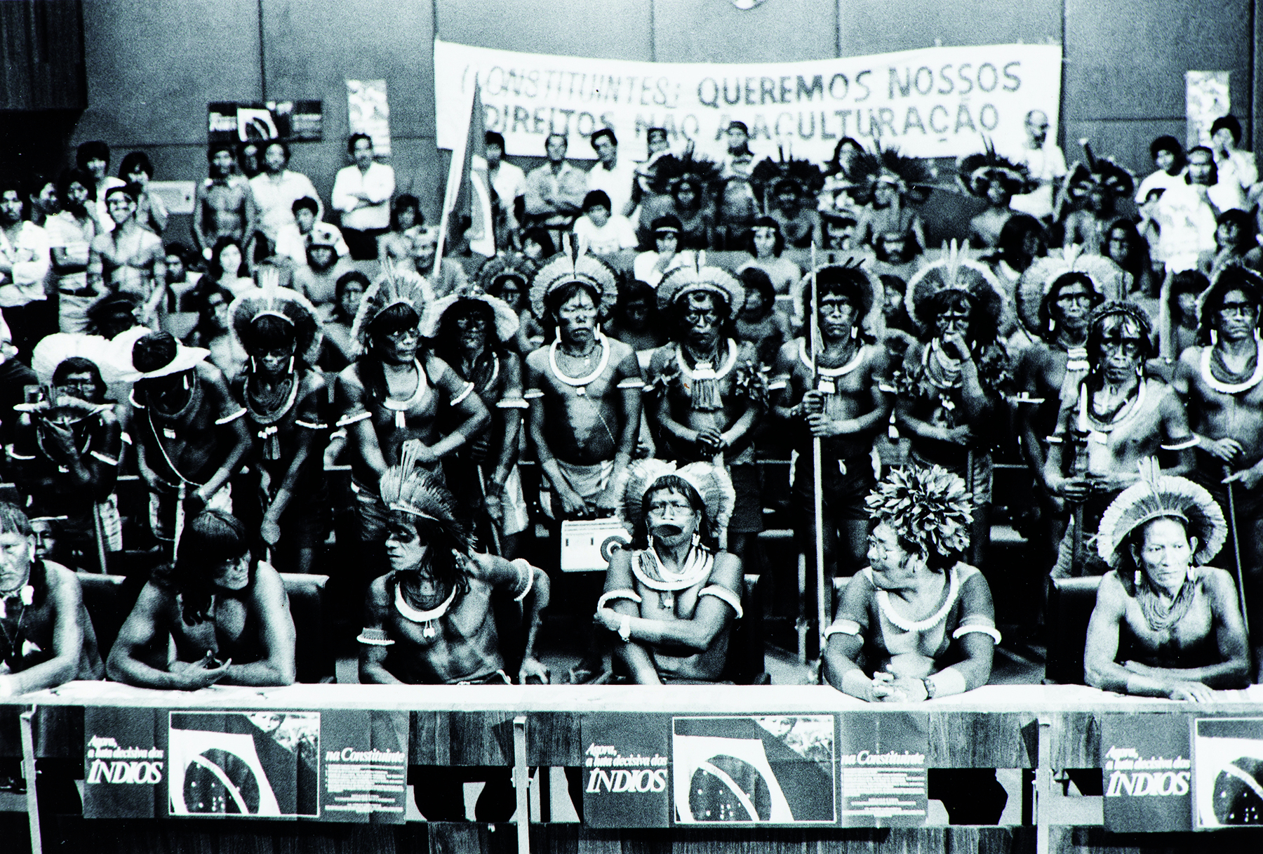 Fotografia em preto e branco. Vários indígenas reunidos em um espaço fechado. Em primeiro plano, alguns deles estão sentados atrás de uma bancada que leva a bandeira do Brasil. Ao fundo muitos indígenas em pé, de frente para o ângulo da fotografia. Ao fundo, uma faixa com os dizeres: 'CONSTITUINTES: QUEREMOS NOSSOS DIREITOS. NÃO À ACULTURAÇÃO'.