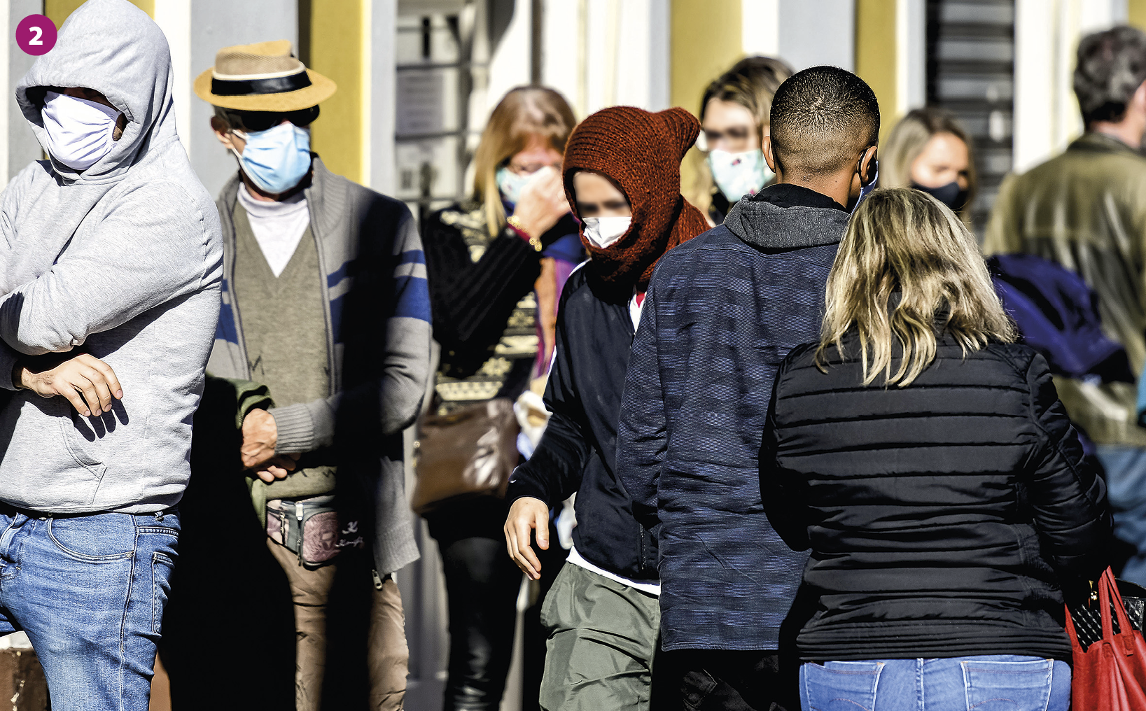 Fotografia 2. Pessoas agasalhadas com jaquetas grossas e toucas, utilizando máscaras de proteção no nariz e boca, trafegam por uma via em um local aberto. Há a presença da luz do sol.