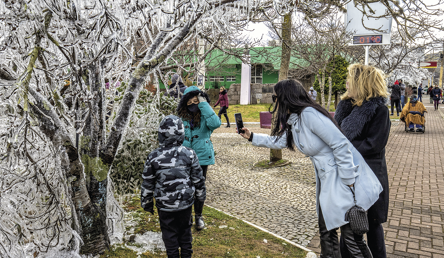 Fotografia. Pessoas estão agasalhadas em uma praça. Em destaque, duas crianças se encontram ao lado de uma árvore coberta por gelo, e uma mulher as fotografa usando um celular. Ao fundo, outras árvores encobertas por gelo e um grande relógio termômetro de rua indicando que a temperatura no local é de um grau Celsius.