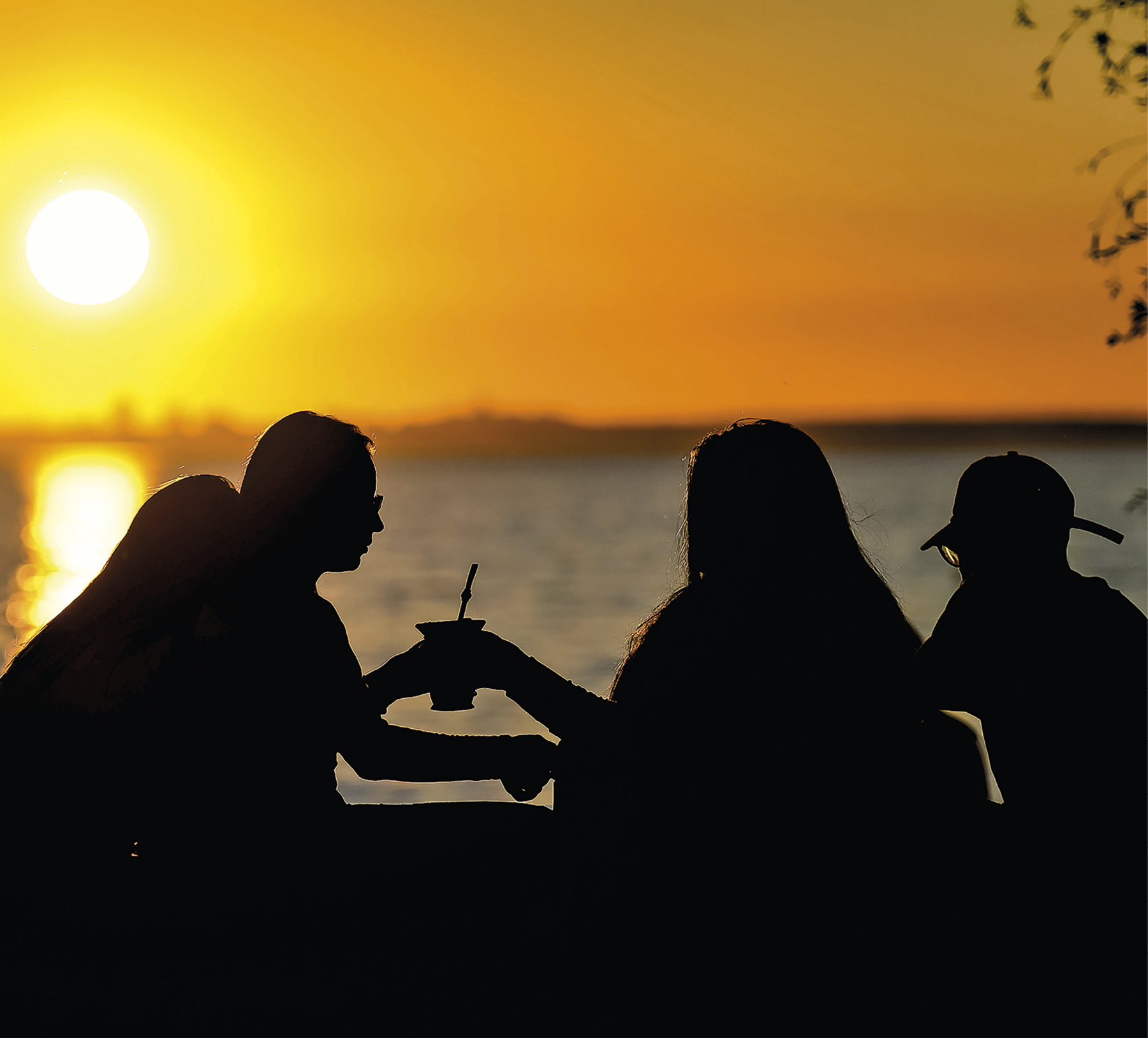 Fotografia. No primeiro plano, silhueta de quatro pessoas sentadas e duas delas seguram um chimarrão. Ao fundo, sol e pôr-do-sol refletido em uma grande porção de água.
