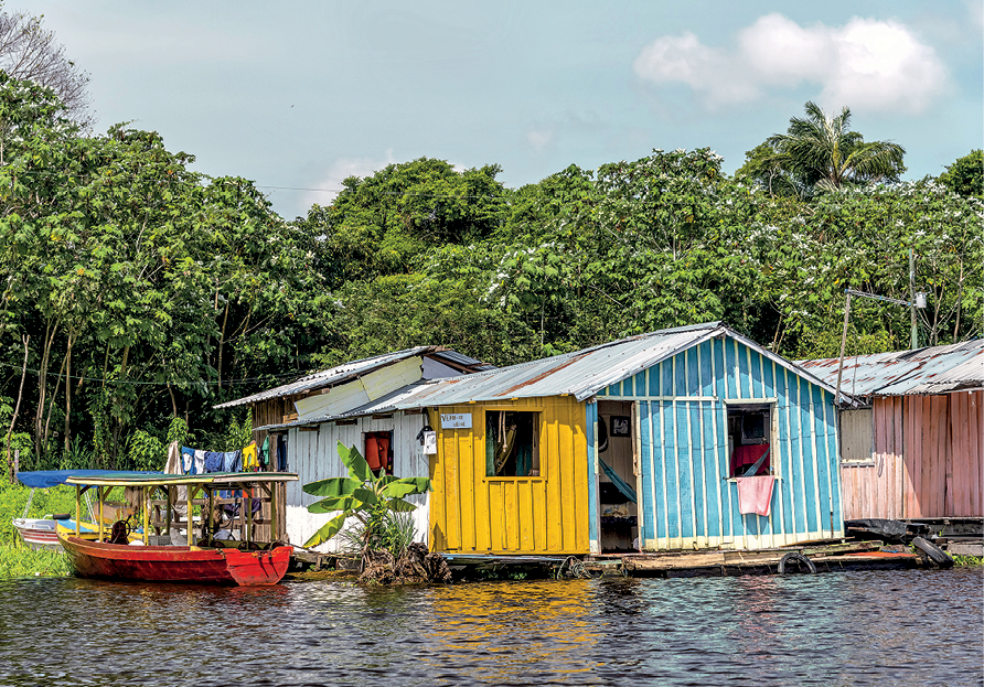 Fotografia. Imagem de casas de madeira coloridas construídas sobre as águas escuras de um rio. À esquerda, pequena embarcação. Ao fundo, vegetação verde e abundante.